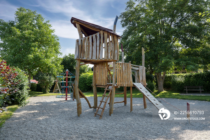 Spielhaus aus Holz auf einem Kinderspielplatz mit Fallschutzkies in einem kleinen schattigen Park mi