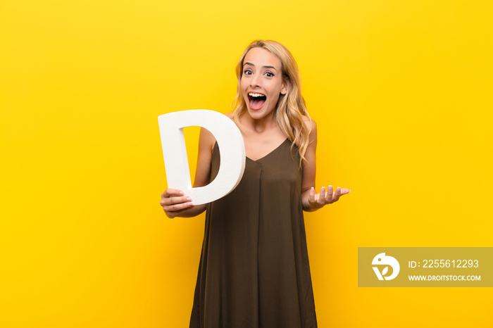 young blonde woman surprised, shocked, amazed, holding the letter D of the alphabet to form a word o