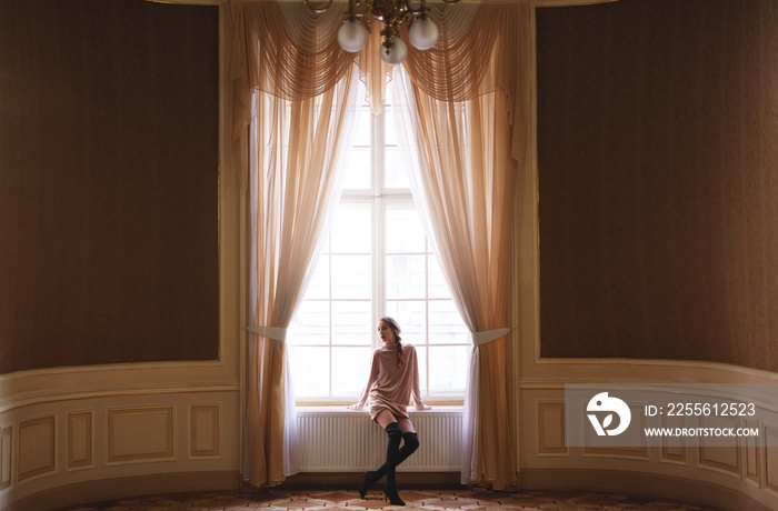 pretty elegant woman sitting at the big window and posing in the big wide palace hall