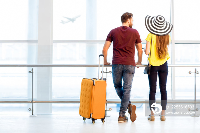 Young couple in colorful t-shirts waiting with baggage near the window at the departure area of the 