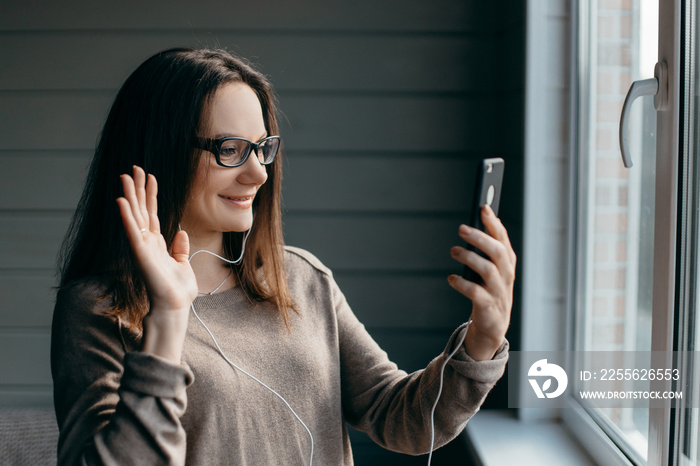 Happy brunette woman in glasses making facetime video calling with smartphone at home, using zoom me