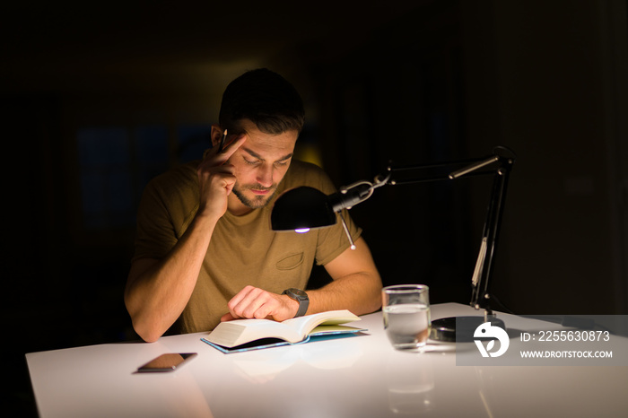 Young handsome man studying at home, reading a book at night