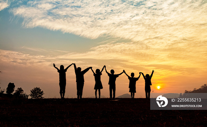 Silhouette of happy children standing with raised hands on the mountain at the sunset time.