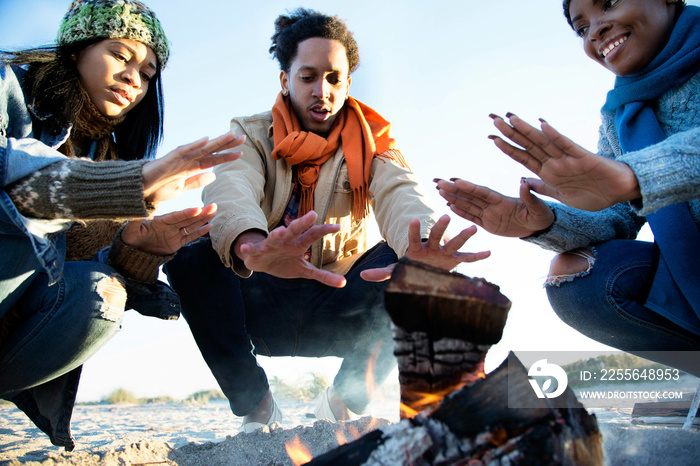 Three friends on beach, warming hands over camp fire