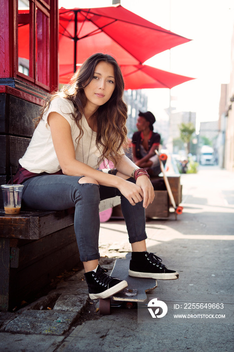 Young female sitting on street bench with skateboard build structure and red sunshades in background