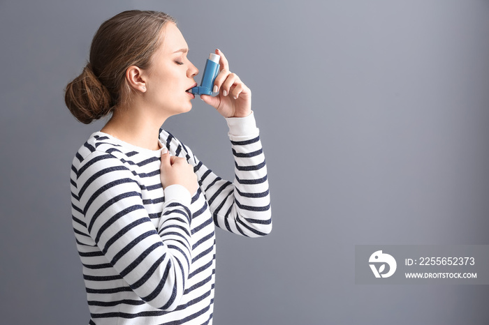 Young woman with inhaler having asthma attack on grey background