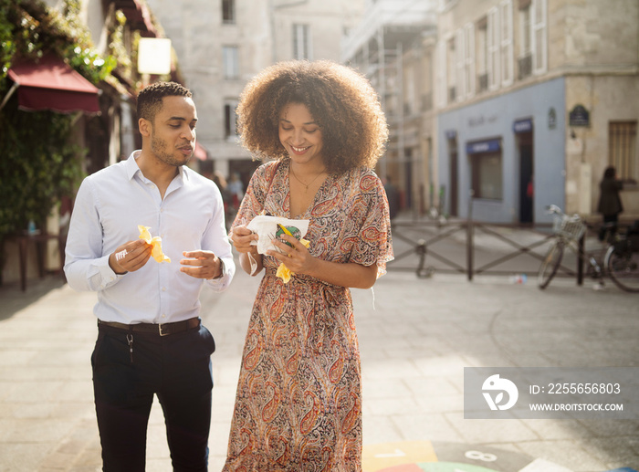 Happy couple eating food while standing on street against buildings in city