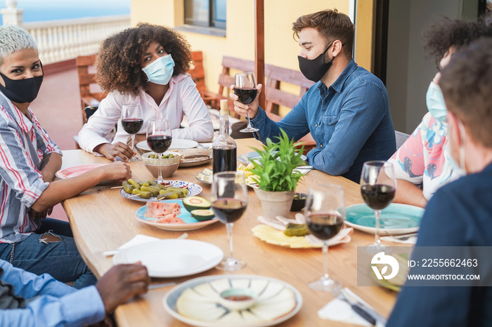 Multiracial friends eating and drinking wine together at home with protective masks - Social distanc