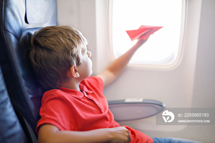 Little kid boy playing with red paper plane during flight on airplane