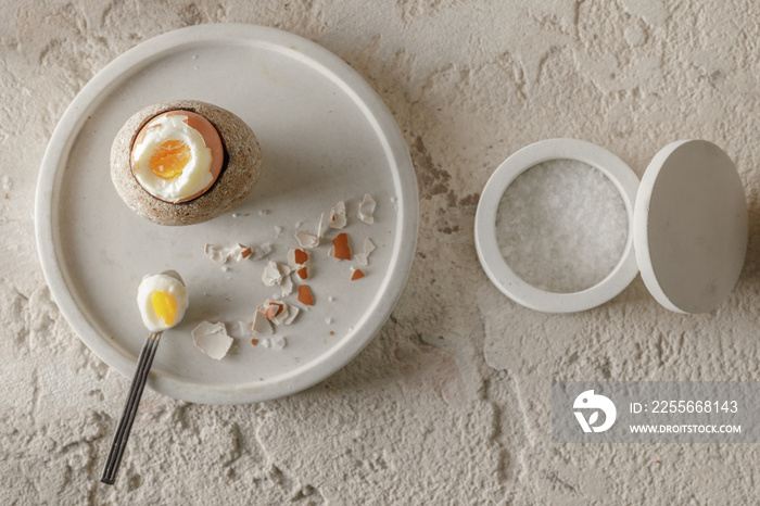 一个煮熟的鸡蛋放在石墙鸡蛋杯里，鸡蛋壳碎片放在混凝土托盘里，盐片放在wh里