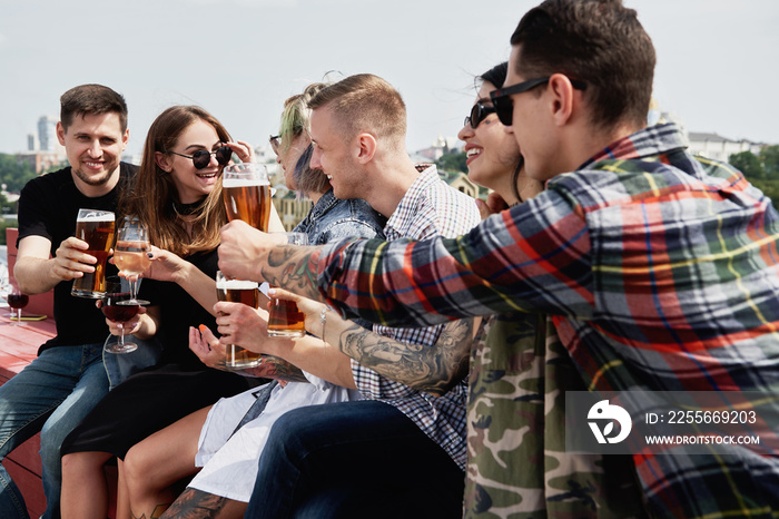 Young happy friends drinking beer before festival at outdoors pub on roof, toasting and laughing, co