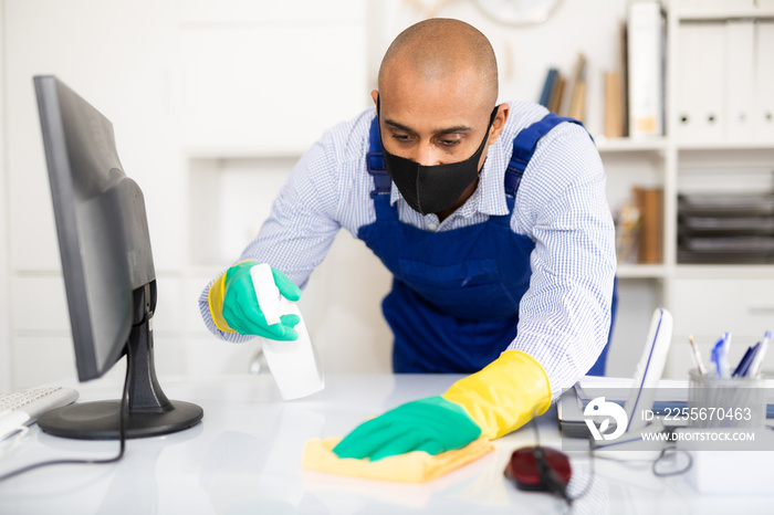 Man professional cleaner in medical mask cleaning desk with computer in office