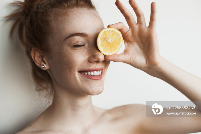 Ginger caucasian woman with freckles is covering her eye with a lemon and smile on a white studio wa