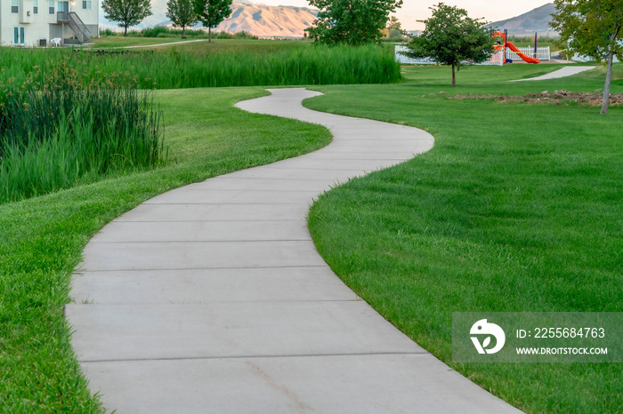 Narrow paved pathway winding through lush green grasses of park with playground