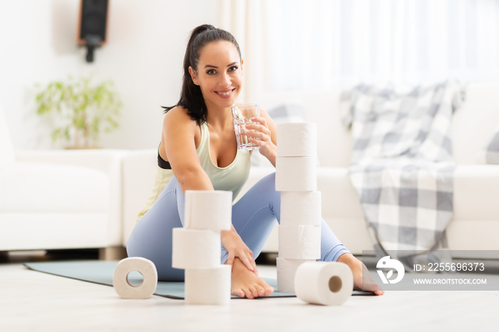 Smiling woman drinks a glass of water aftera home workout surrounded by toilet paper rolls in her li