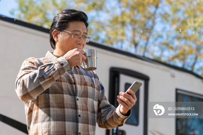 一个老年男人站在房车前看着手机视频喝咖啡