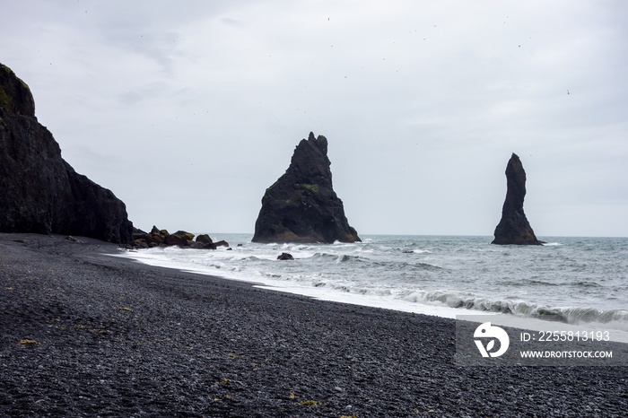 Reynisdrangar basalt rock formation in the Atlantic ocean with black sand beach and rocky mountain in overcast day with grey sky typical for Iceland