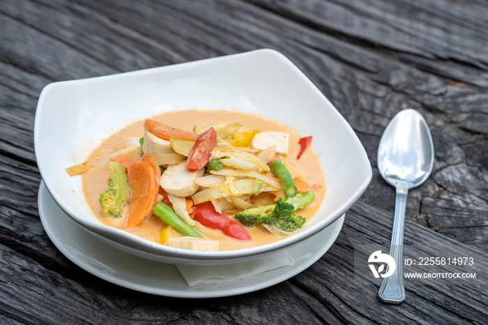 Mixed vegetables in creamy red curry coconut sauce with tofu in a white plate on a wooden table , traditional Thai food.