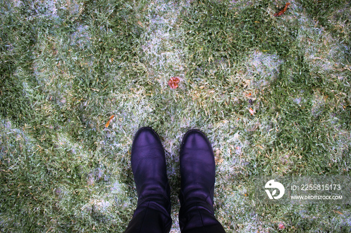 Legs in boots against the background of frozen lawn covered with ice with green grass.