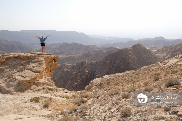 Silhouette of standing woman with arms raised  on rock at beautiful mountain viewpoint. Views of mountain range from Tafilah Highway, Dead Sea depression in the background. Jordan.