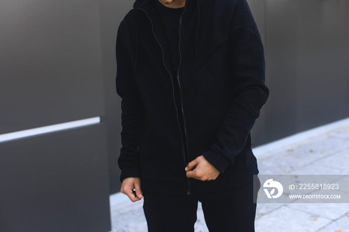 Young man zipping up his hoodie. Man dressing black sport clothes. Grey urban background.