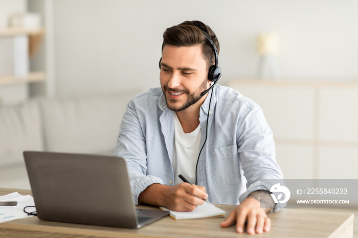 New knowledge online at home. Young man in headphones making notes and looking at laptop computer in