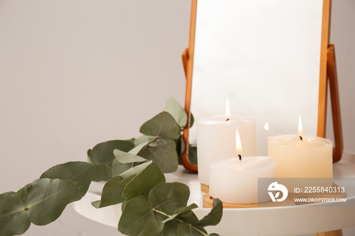Burning candles, eucalyptus leaves and mirror on end table near grey wall