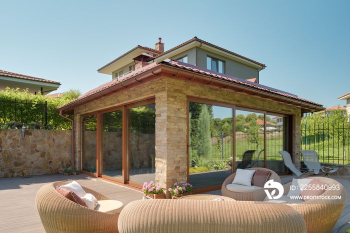 Modern summer house made of glass and stone. Wicker garden furniture on a wooden terrace.