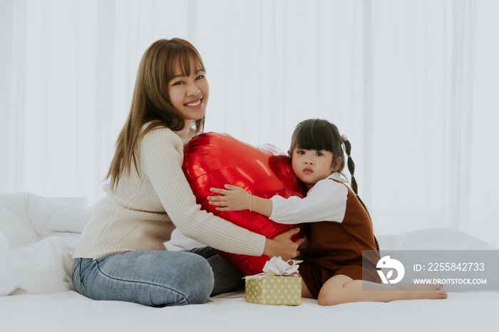 Young asian mom smiling holding heart balloon playing with little 4years old kid daughter in bed roo