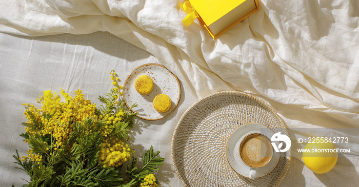 Branches of yellow mimosa flowers on white linen bed sheets, cup or coffee, french yellow macaroons,