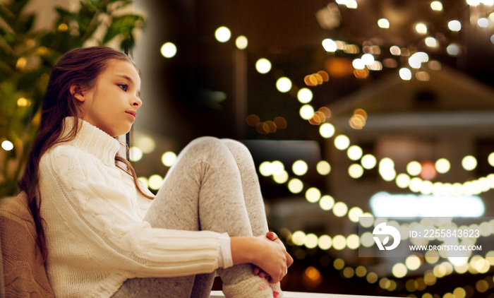 childhood, sadness and people concept - sad beautiful girl in sweater sitting on sill at home window