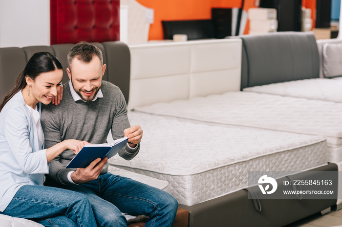 smiling couple with notebook sitting on mattress in furniture store
