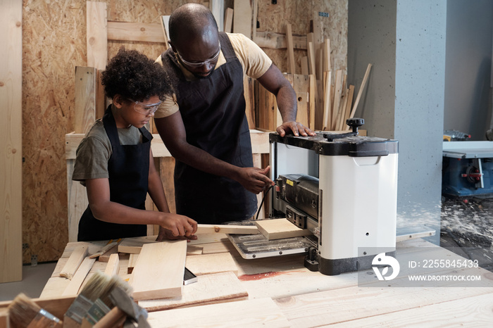 Portrait of African-American father and son cutting wood while working together in carpentry worksho