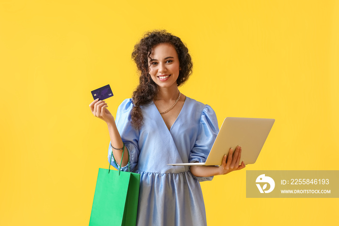 Young African-American woman with credit card, laptop and shopping bag on yellow background