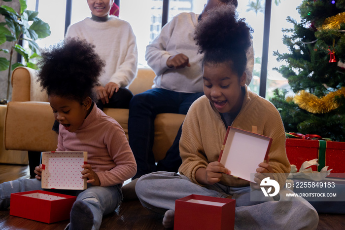 Excited two cute little african american daughters sitting on wooden floor opening christmas present