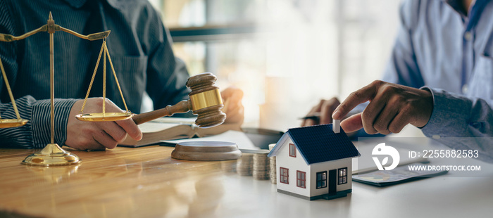 Judge holding wooden judge hammer against house background and businessman documents and house model on wooden table Real estate disputes and property auction concept