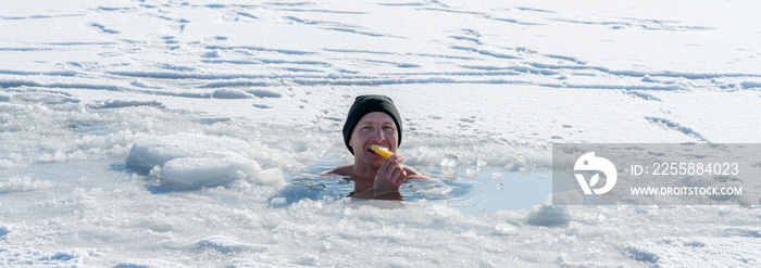 Man sitting in an ice hole while eating ice cream in winter