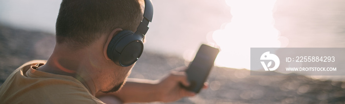 Portrait of a young man sitting on the seashore wearing headphones with a phone in his hands at sunset.