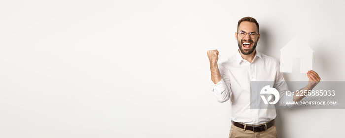 Real estate. Satisfied man rejoicing of founding perfect home apartment, holding paper house model, standing over white background