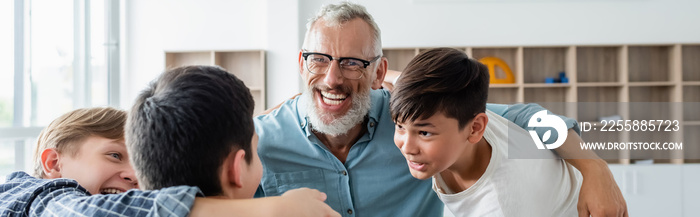 multicultural boys and cheerful middle aged teacher embracing in classroom, banner