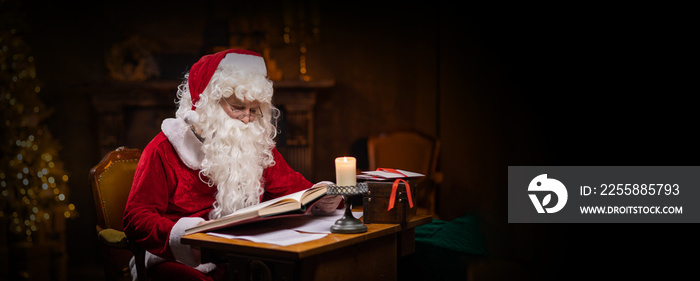 Workplace of Santa Claus. Cheerful Santa is reading the book of wishes while sitting at the table. Fireplace and Christmas Tree in the background. Christmas concept.