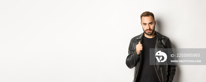 Handsome bearded man expressing confidence, touching his leather jacket and looking self-assured, white background