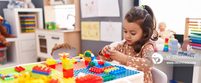 Adorable hispanic girl playing with construction blocks sitting on table at kindergarten