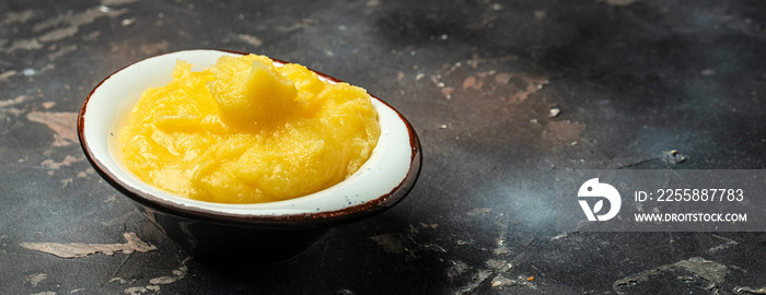 Pure OR Desi Ghee also known as clarified liquid butter in a bowl with wooden spoon on a dark background. Long banner format