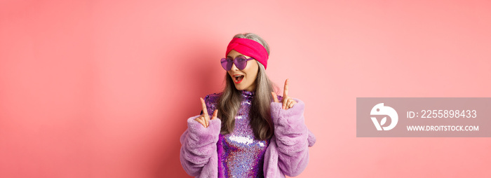 Hipster granny in sunglasses and glittering dress posing for photo with peace, victory signs, standing sassy against pink background