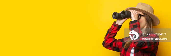 Young woman in hat and plaid shirt looking through binoculars on yellow background