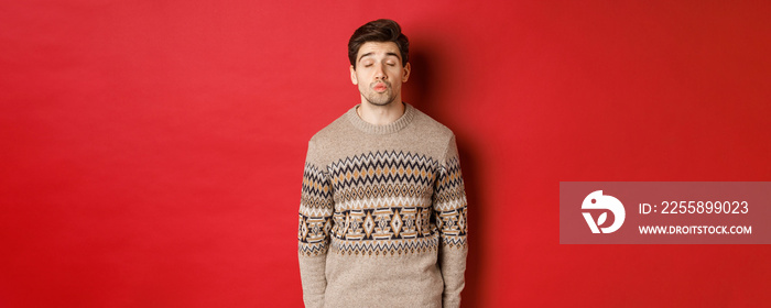 Image of handsome man in christmas sweater pucker lips and close eyes, waiting for kiss under mistletoe, standing over red background