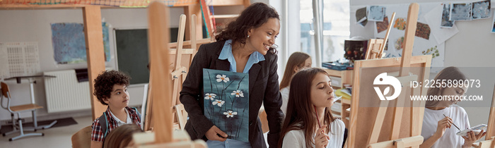 Young beautiful confident teacher is helping a kid to draw on a group lesson in a white modern minimalistic classroom.