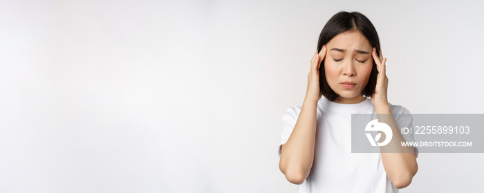 Portrait of asian girl feeling headache, migraine or being ill, standing in white t-shirt over white background