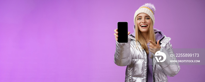 Satisfied amused good-looking blond girl suggest take look smartphone display smiling happily pointing mobile phone delighted talking about awesome new app features, standing purple background